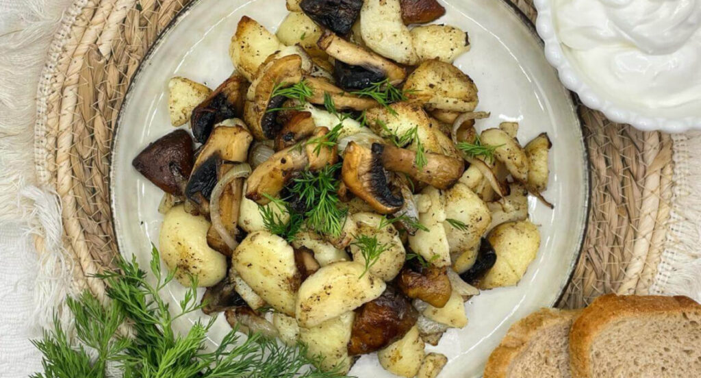 Жареная картошка с грибами на белой тарелке с кусочками зелени и сметаной